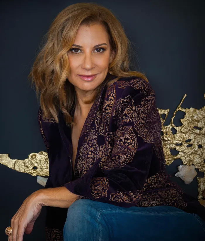 Portrait of Janet Morais, Founder and CEO of ByKOKET Group, wearing a purple and gold patterned jacket, with the sculptural brass details of KOKET’s Empress Console visible behind her.
