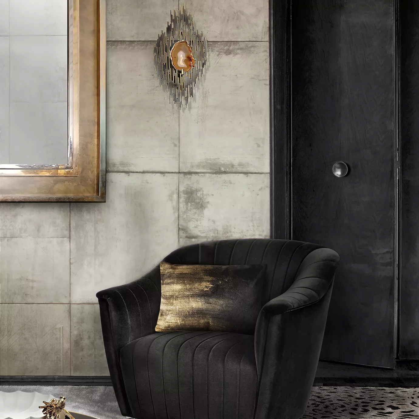 Elegant corner with a armchair upholstered in black velvet with a decorative black and gold-toned pillow, a sculptural golden Vivre Sconce with an illuminated agate center on the grey-toned textured wall, and a mirror with a golden frame.