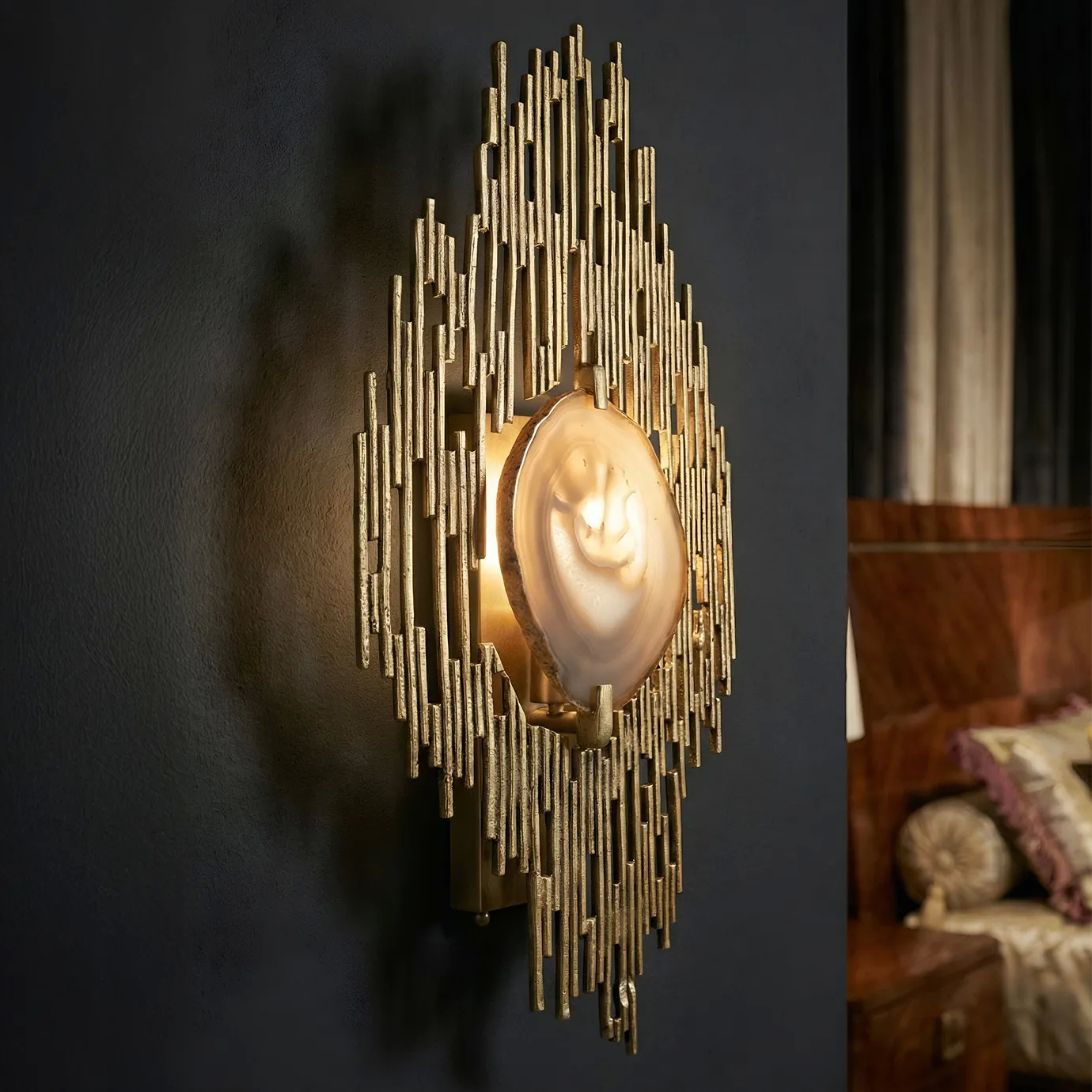 Close-up of the KOKET Vivre wall sconce showcasing its sculptural gold-plated brass frame and polished agate stone center, casting ambient luxury lighting against a dark wall.