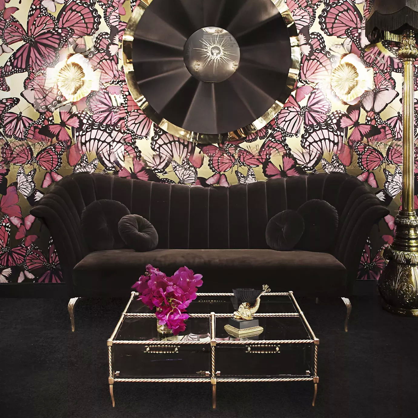 Elegant living room with a Caprichosa Sofa in black velvet with a golden base and matching round cushions, a coffee table with a golden frame and glass top, and dramatic floral and butterfly wallpaper.