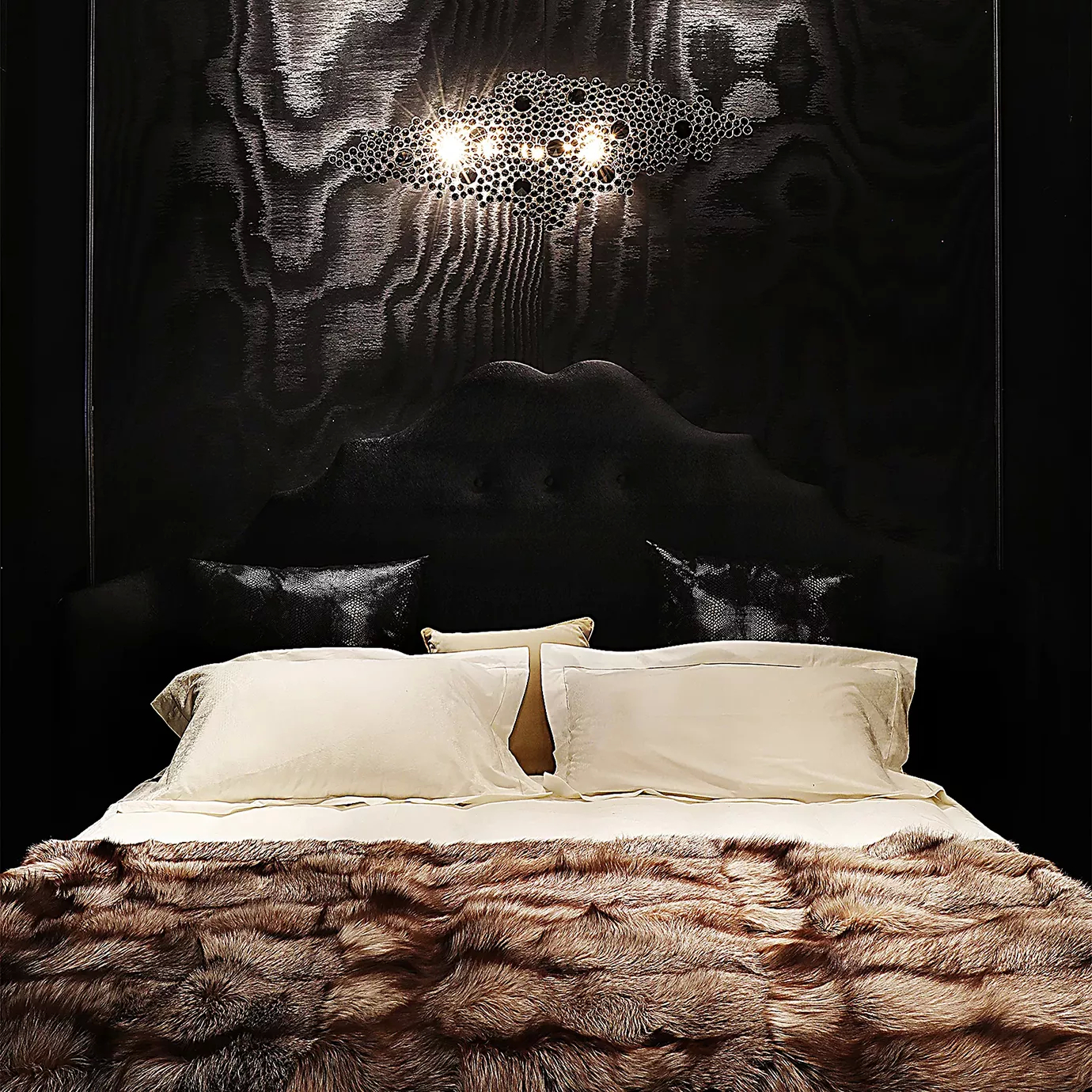 Elegant bedroom with the Alexia Bed featuring a high black velvet headboard under a modern wall lamp and over light bedding with a fur throw.