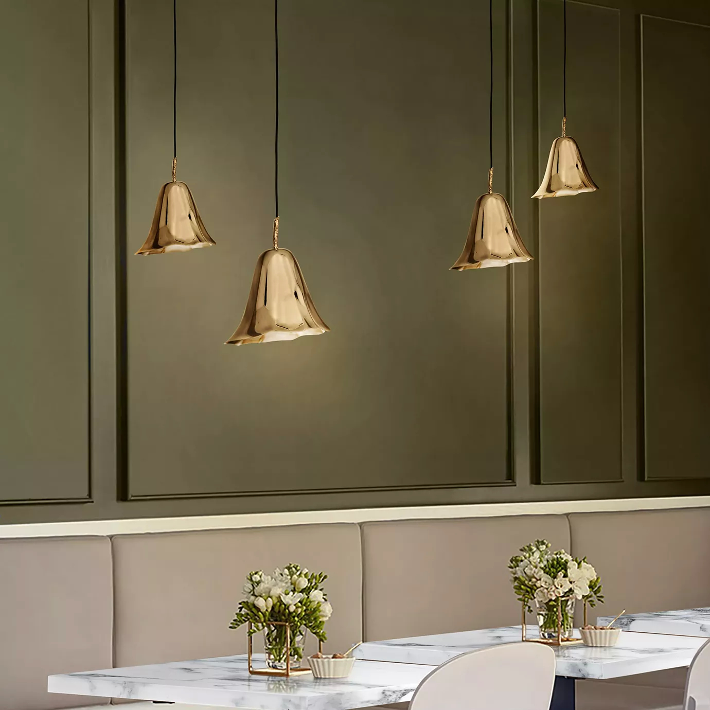 Elegant dining area with four golden Gia Pendants suspended above white marble tables with floral arrangements and light chairs, in front of a dark green wall with embossed details.
