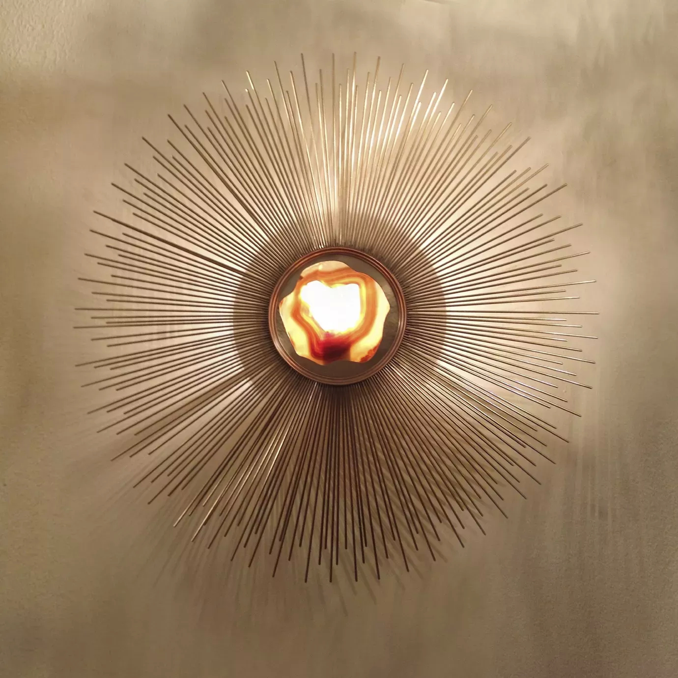 Elegant Brilliance Sconce with a sculptural radial design in golden tones mounted on a light wall, featuring an illuminated translucent central detail with shades of white and orange.
