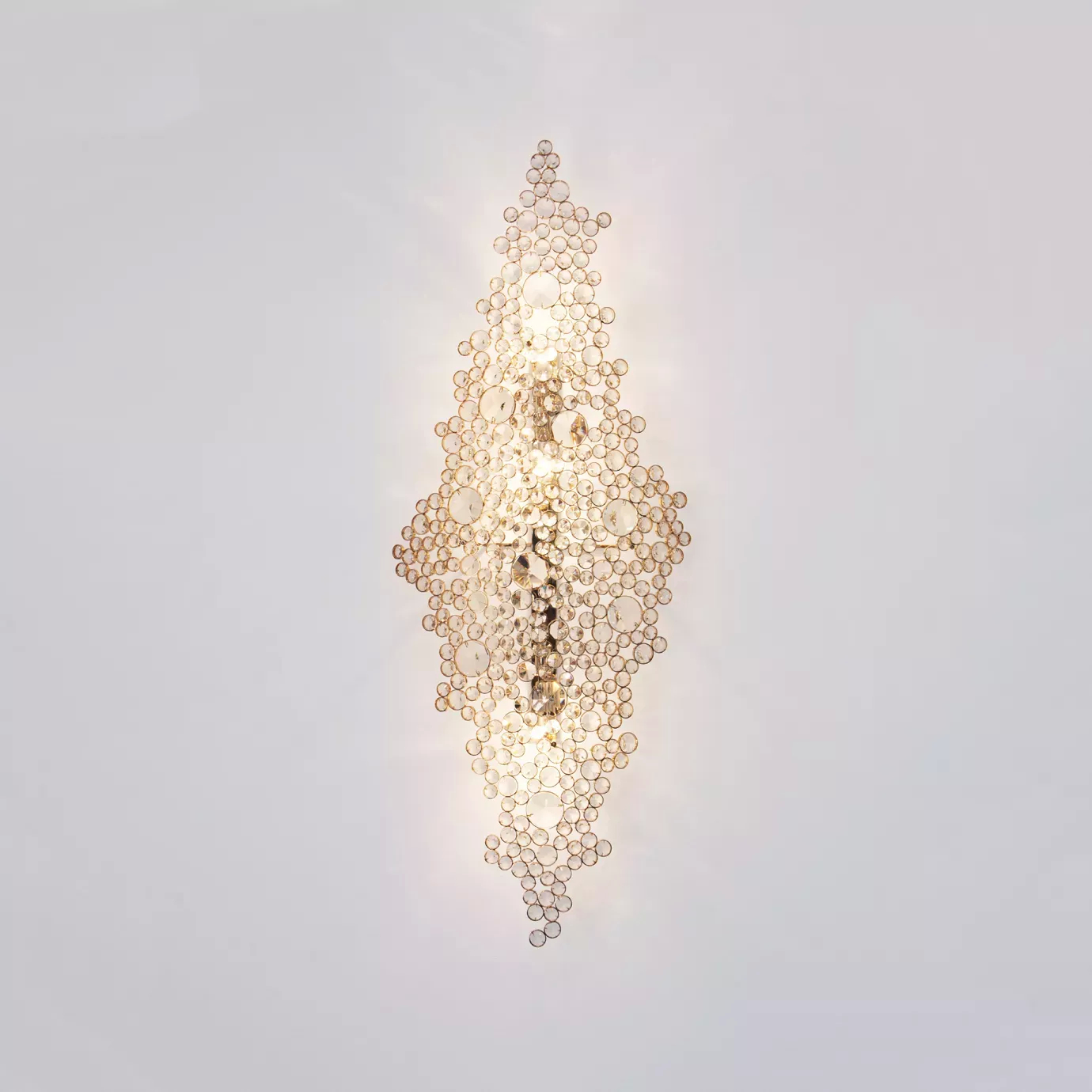 Elegant Eternity Sculpture with a vertical sculptural design adorned with numerous clear crystals of varying sizes on a golden frame, illuminated on a white wall.
