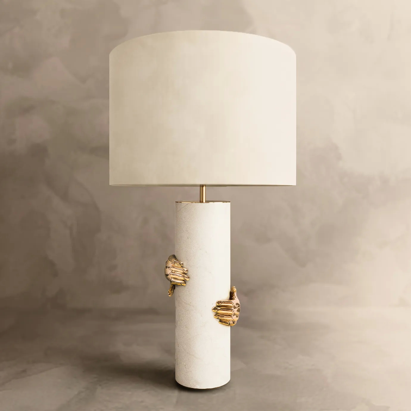 Elegant Vengeance Table Lamp with a white marble cylindrical base adorned with sculptural golden hand details gripping it, and a white lampshade, on a grey surface with a textured background.