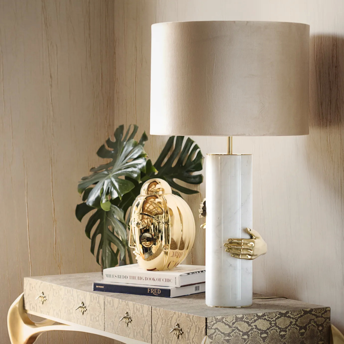 Elegant Vengeance Table Lamp with a white marble cylindrical base adorned with sculptural golden hand details gripping it and a beige lampshade, placed on a textured table next to a golden sculpture and green plants.