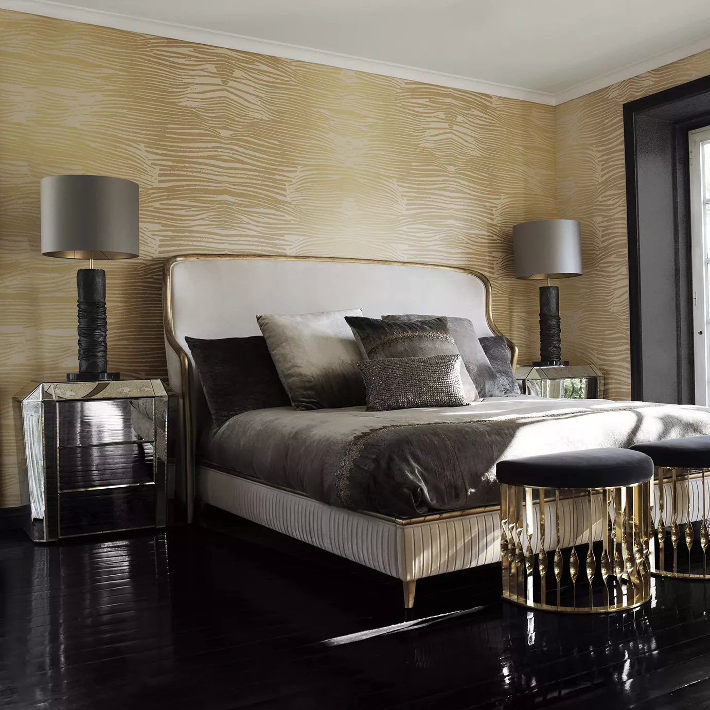Elegant bedroom with a light-toned upholstered bed with golden accents and brown and grey-toned pillows, mirrored nightstands with Ruchê Table Lamps featuring textured brown and gold-toned bases and grey lampshades, and two black ottomans with sculptural golden bases at the foot of the bed, on a dark wooden floor and with beige and gold-toned textured wallpaper.