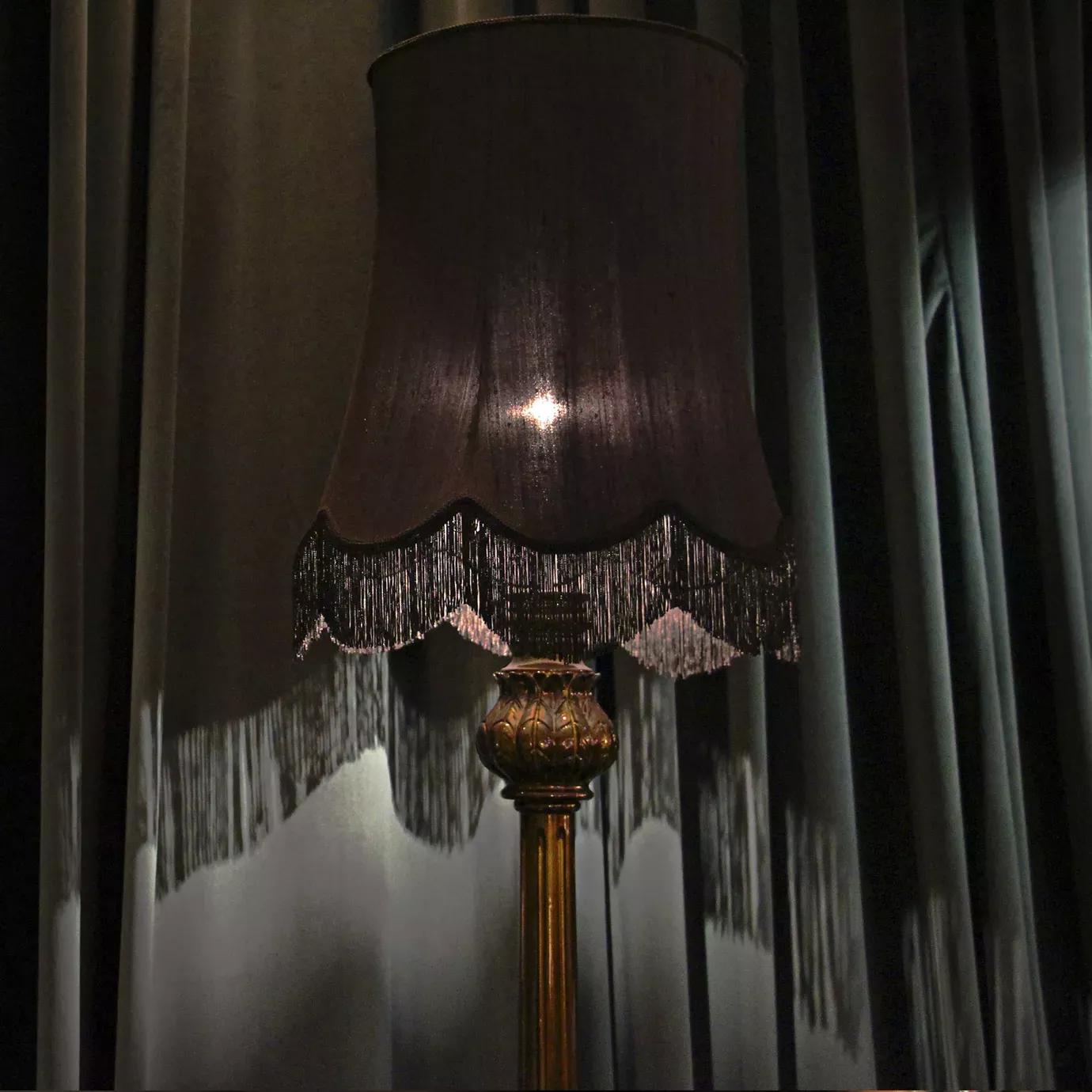 Elegant Lotus Floor Lamp with an ornate sculpted base in golden tones, a decorative vertical stem, and an illuminated black lampshade with fringe details, in front of textured grey and green-toned curtains.