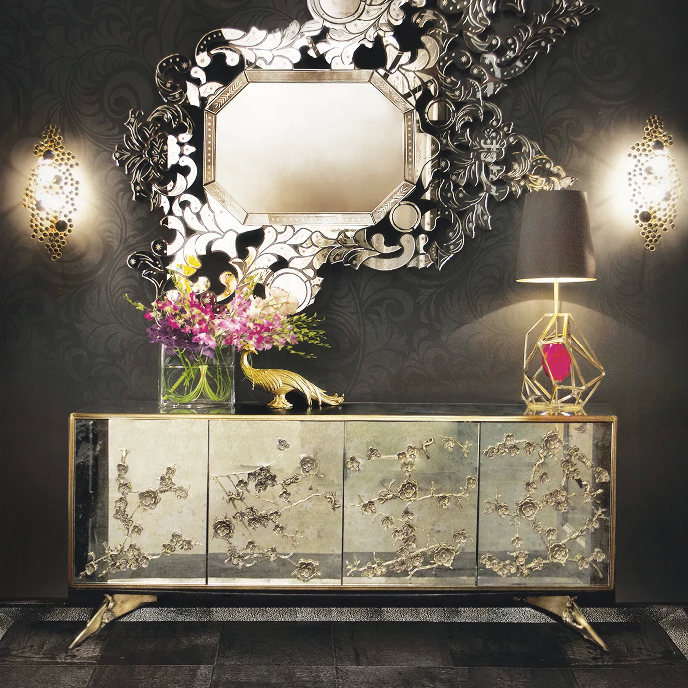 A room in grey hues, featuring an elegant Spellbound Aged Mirror Cabinet, with golden floral details on its four front doors. A silver mirror is set above the cabinet, with two sconces, one on each side.