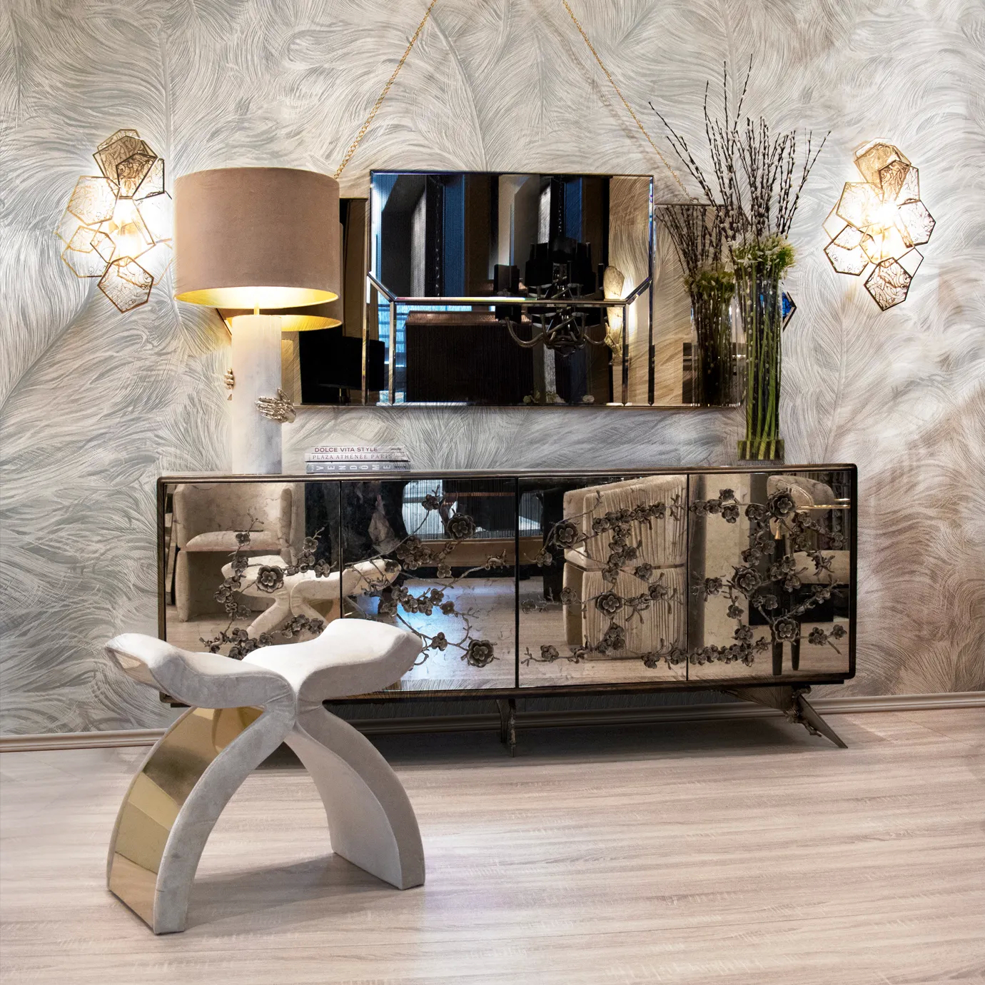 A room in neutral hues, featuring an elegant Spellbound Aged Mirror Cabinet, with golden floral details on its four front doors. There’s a mirror above the cabinet and a creamy bench in front of it. Set on the cabinet, there is a table lamp with golden details.