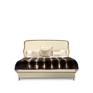 Elegant Forbidden Plain Bed with a headboard and frame upholstered in light beige fabric with golden trim along the edges, adorned with cushions in beige tones and a brown faux fur throw.
