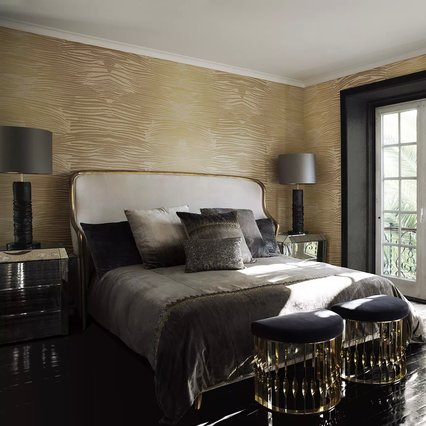 Elegant bedroom with the Forbidden Plain Bed featuring a white fabric upholstered headboard and frame with golden accents, adorned with cushions in grey tones and a grey velvet throw, flanked by glossy black nightstands with marble tops and Table Lamps with textured black bases and grey lampshades, and two black ottomans with golden details and fringes at the foot of the bed, on a glossy dark wooden floor and against walls with beige and gold-toned textured wallpaper.