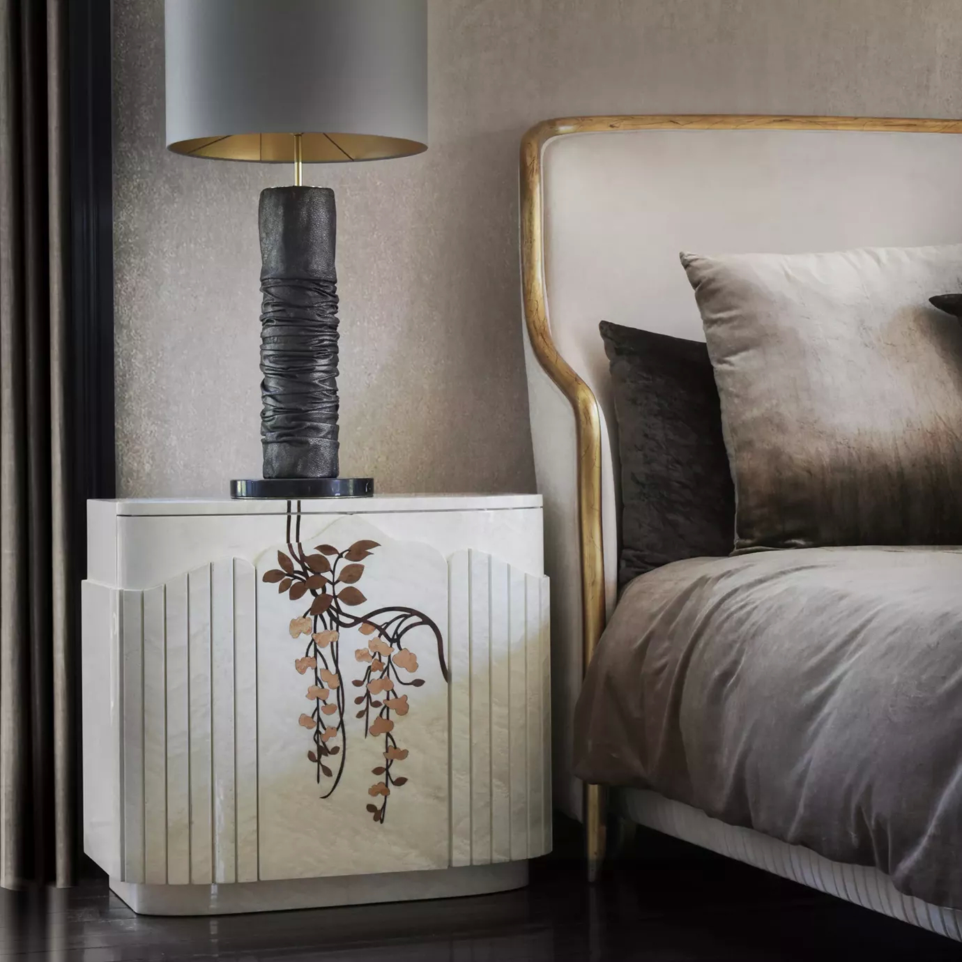 Elegant bedroom corner featuring the cream-white Fleur Nightstand adorned with a brown-toned floral pattern, topped with a table lamp with a textured black base and a grey lampshade, next to a bed with a cream-toned upholstered headboard with golden accents and grey-toned bedding, on a dark floor.
