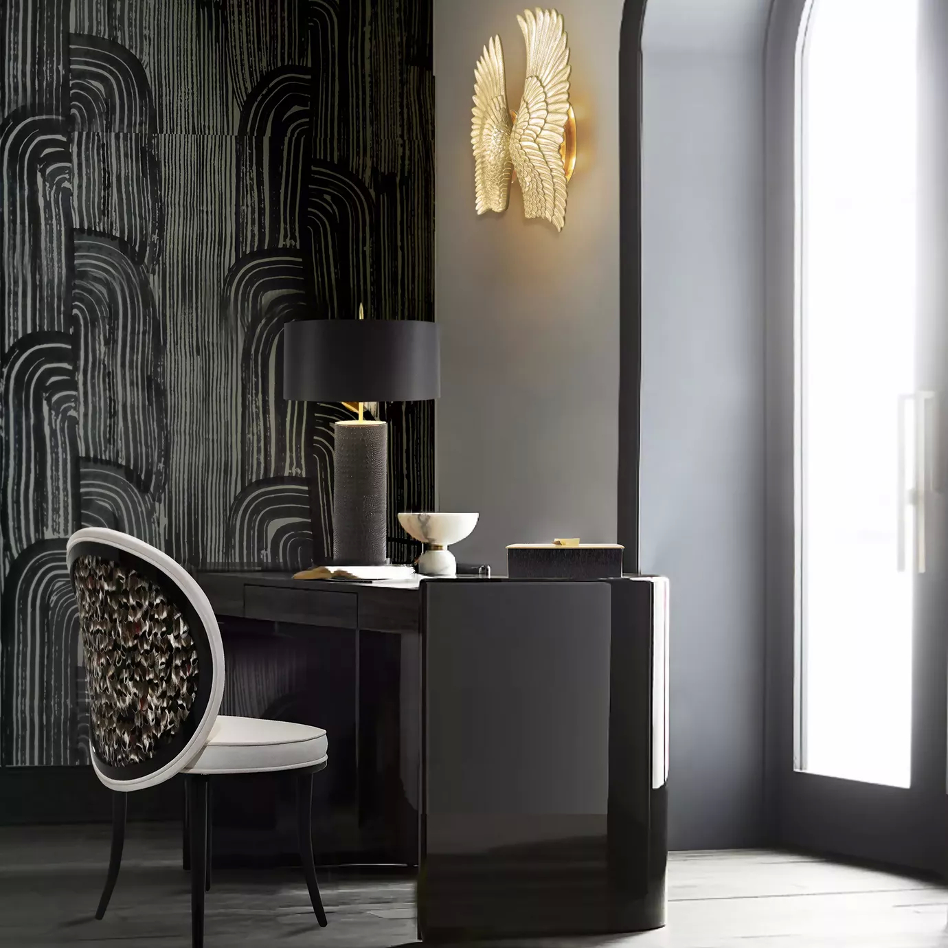 Elegant office corner featuring the Maleeha Dining Chair with a backrest adorned with a feather pattern in shades of brown and white, white upholstery, and tapered black legs, placed next to a dark desk with a table lamp featuring a textured dark base and a black lampshade, and several decorative objects, against a wall with black and white textured wallpaper and a golden wing-shaped sconce.