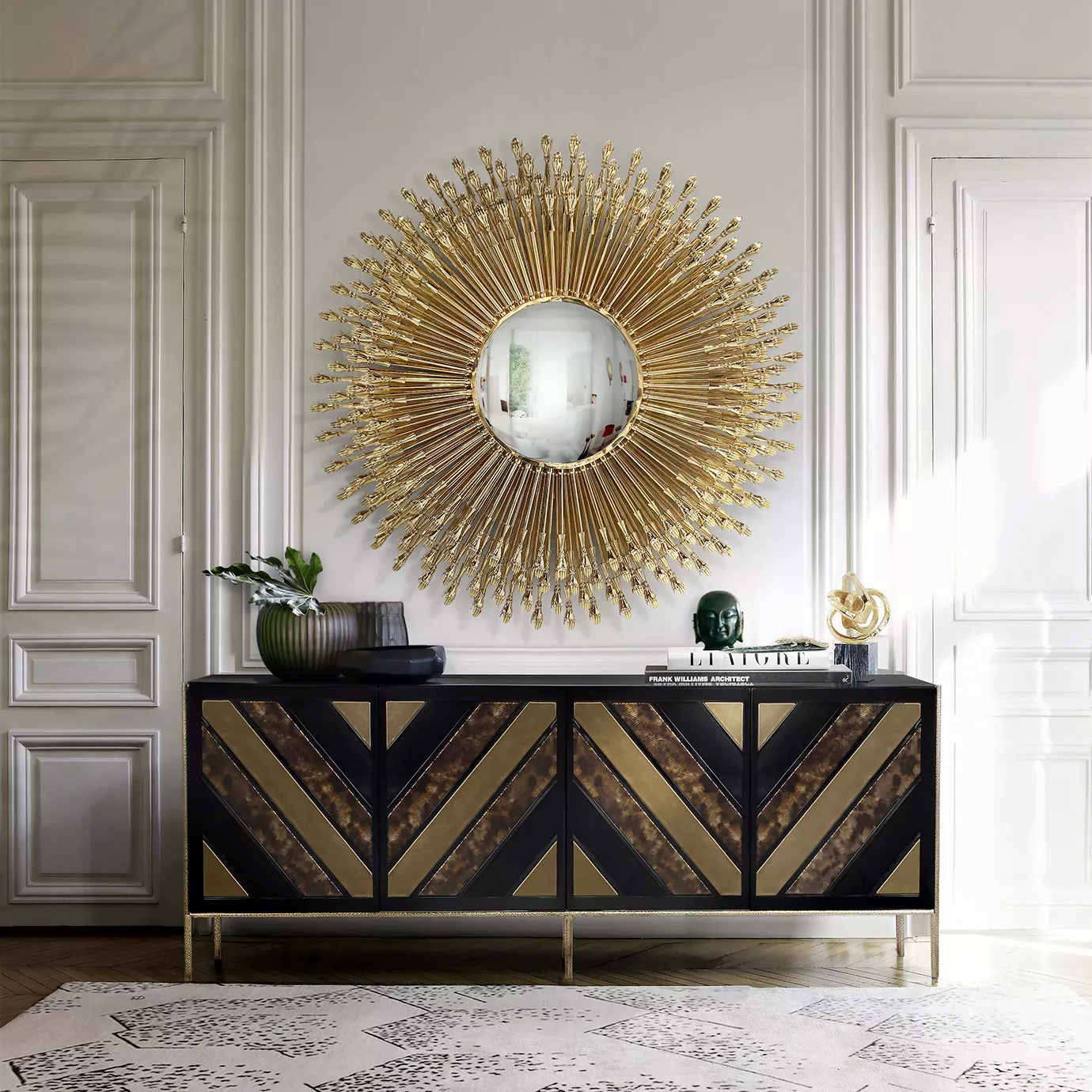 Neutral room with a cabinet in brown and black hues and some decorative elements on top. Above the cabinet there is a Goddess Mirror, showcasing its golden frame portraying multiple tiny hands around it.