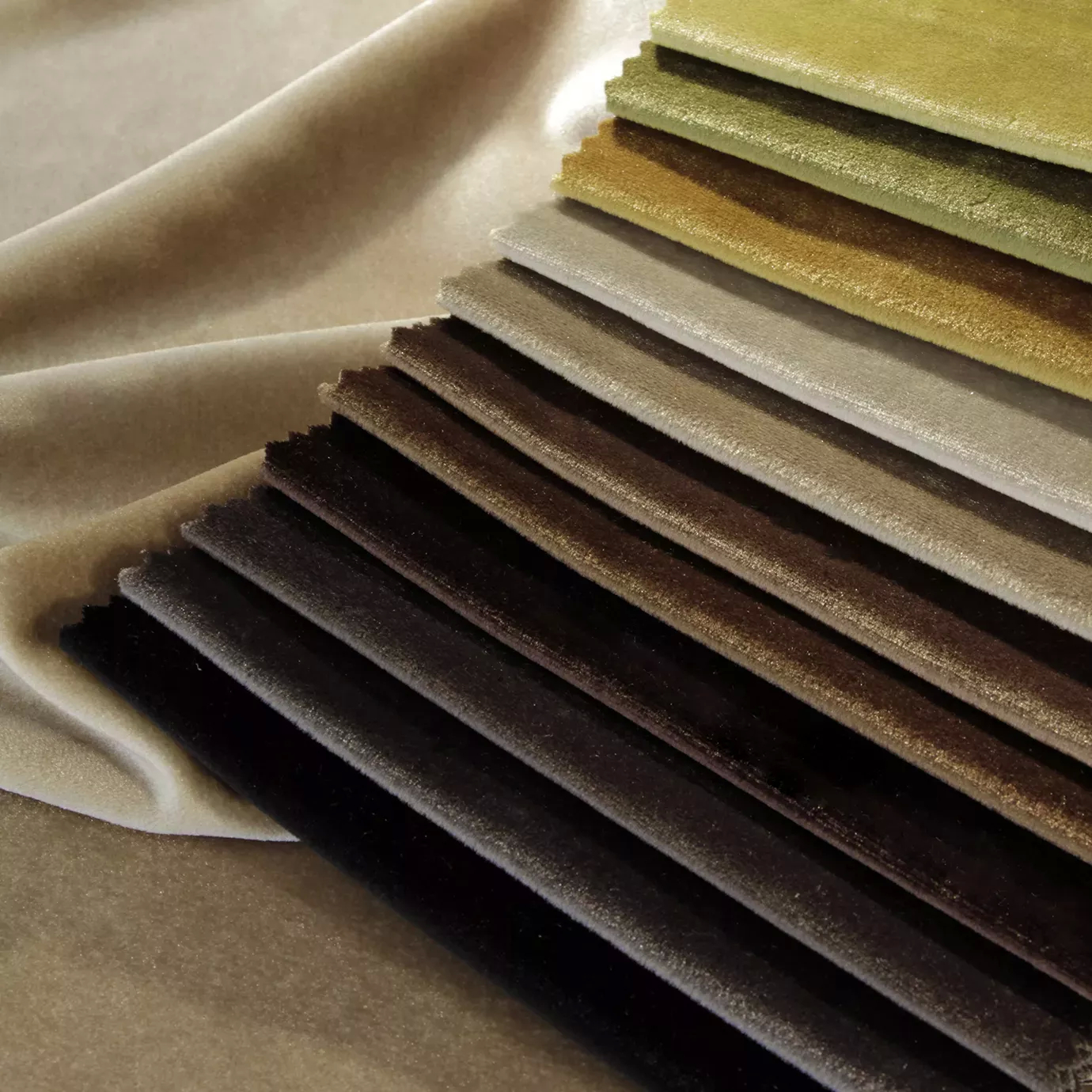 Elegant presentation of a variety of Silky Velvet fabric swatches in diverse soft and luminous tones, layered on a flowing beige fabric, showcasing the smooth texture and silky sheen of the collection.