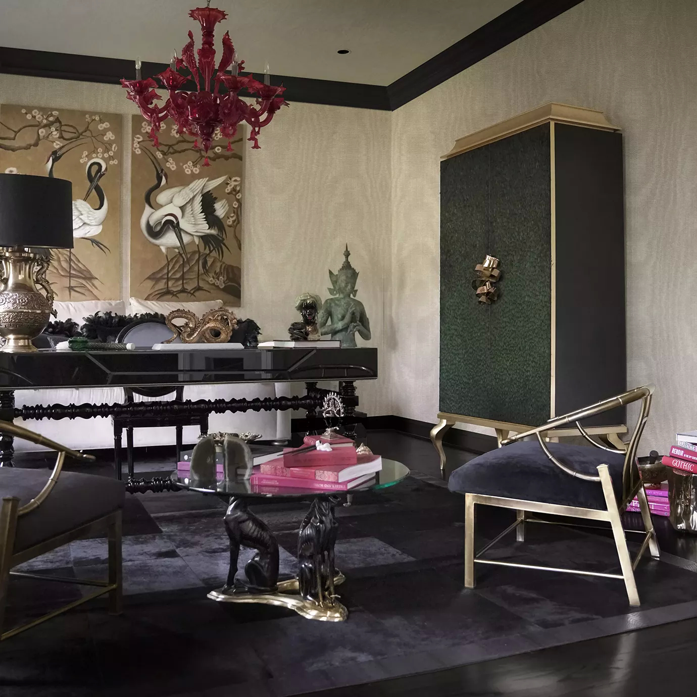 Luxurious living room, in black and golden hues. The Divine Peacock Feather Armoire, covered in delicate dark green peacock feathers, with a golden frame and adorned with a golden handle resembling a ribbon, is set on the corner of the room. There are various black and golden armchairs and a black desk set on a black rug. The room has a also a few pops of pink, on the chandelier and on the books set on the center table.