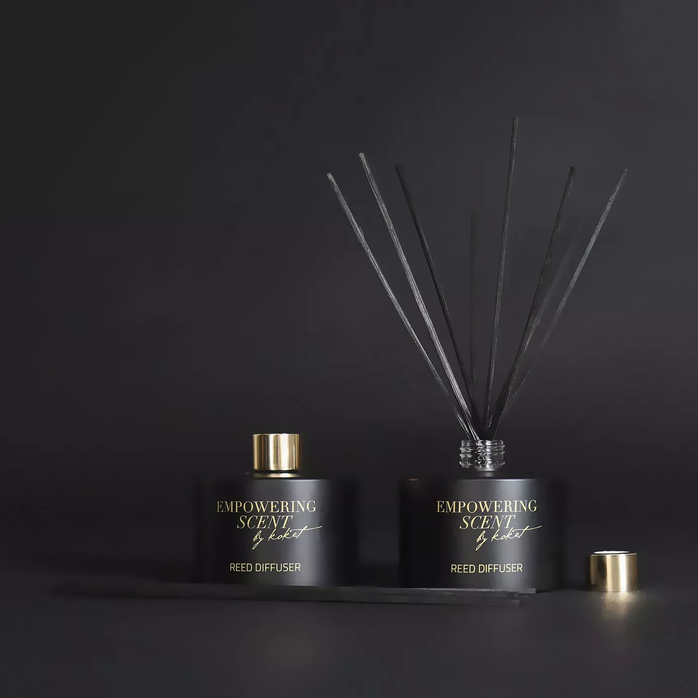Two Empowering Diffusers on a dark surface: black bottles with gold accents and reeds, one assembled, the other with the cap off.