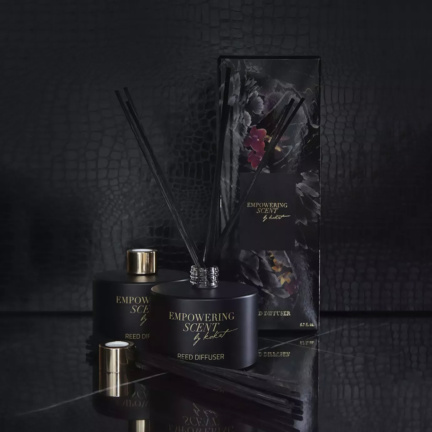Two Empowering Diffusers with reeds, one with a gold cap, next to a floral-patterned box on a dark surface.