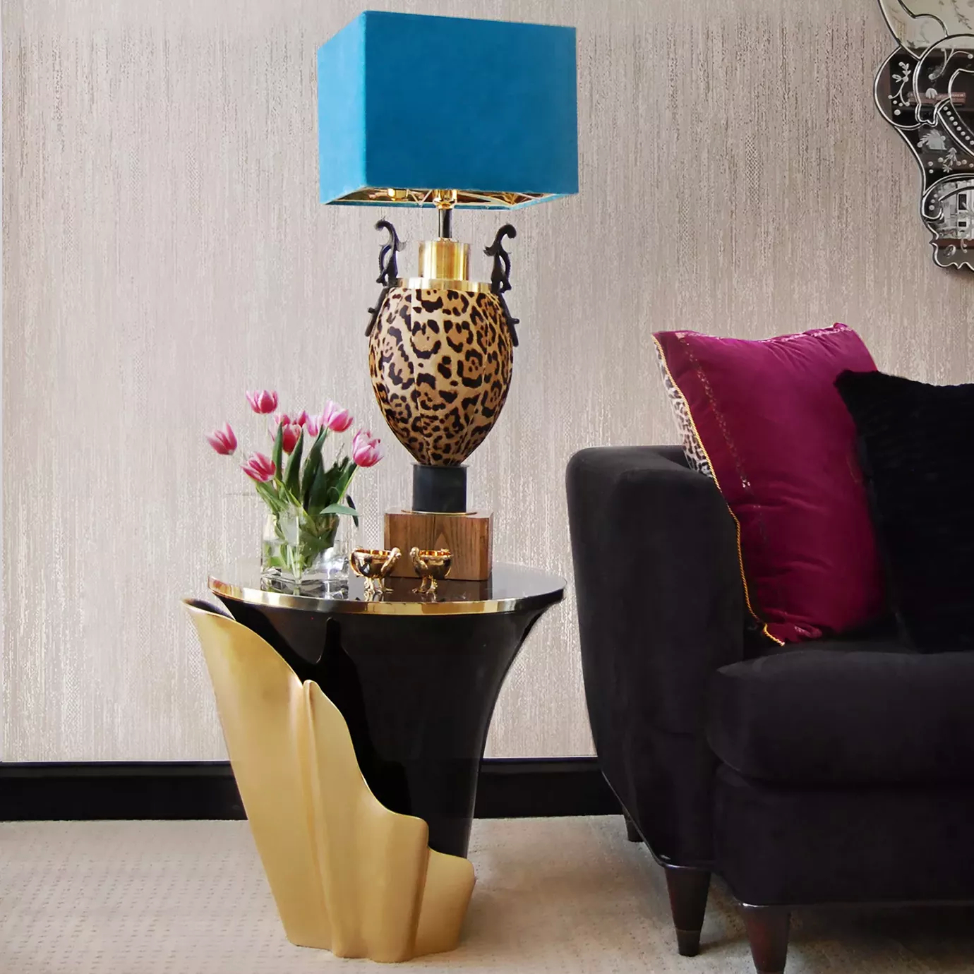 A neutral colored space, featuring a Yasmine Side Table. The side table has a black marble tabletop with golden edges, set on a solid wooden black base, adorned with a golden structure that arches back and forth. There is a blue table lamp on top of the side table and next to it there is a black sofa, with a fushcia pillow.