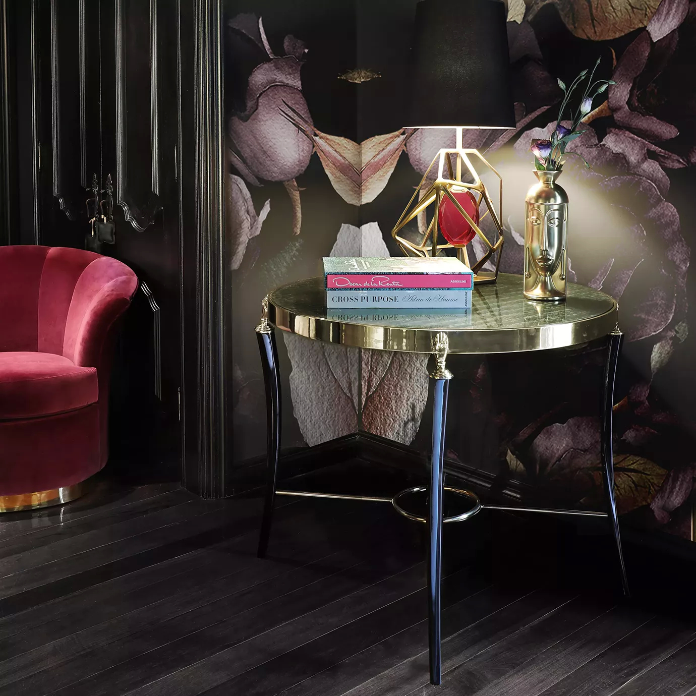 Gisele Table in a luxurious room with a dark floral wallpaper, a red velvet chair, a golden lamp, books, and a vase.