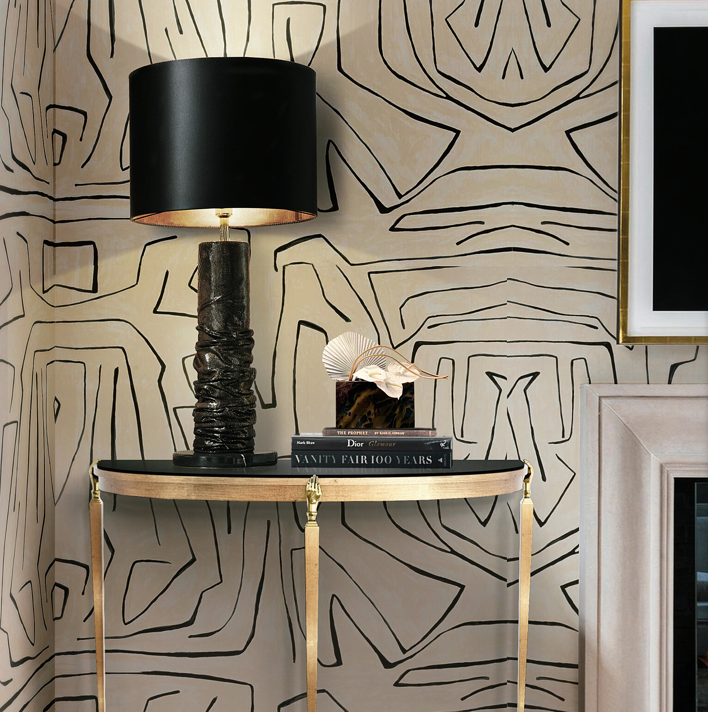 The Gisele Demi Lune Console, with its black top and gold legs, stylishly holds a lamp and books against patterned wallpaper.