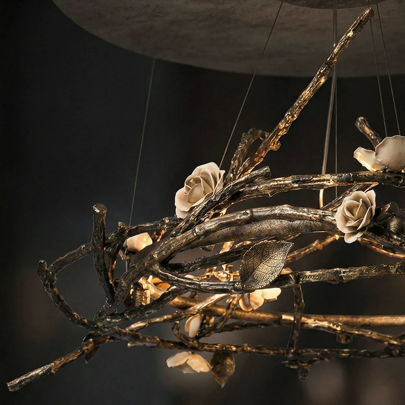 Rose Round Chandelier in aged brass with sculptural branches, metallic leaves, and cream rose-shaped accents, suspended by thin wires in a modern interior.