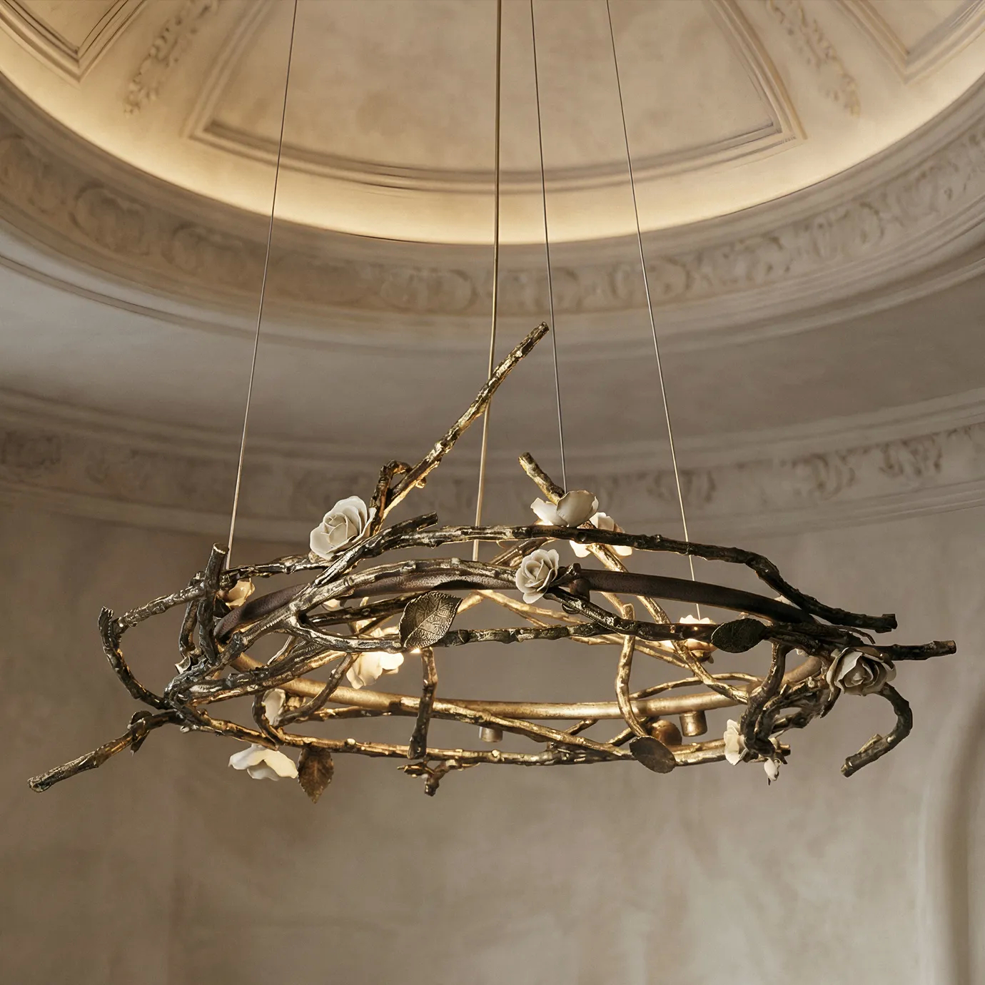 Rose Round Chandelier in aged brass with sculptural branches, metallic leaves, and cream rose-shaped accents, suspended by thin wires in a modern interior.