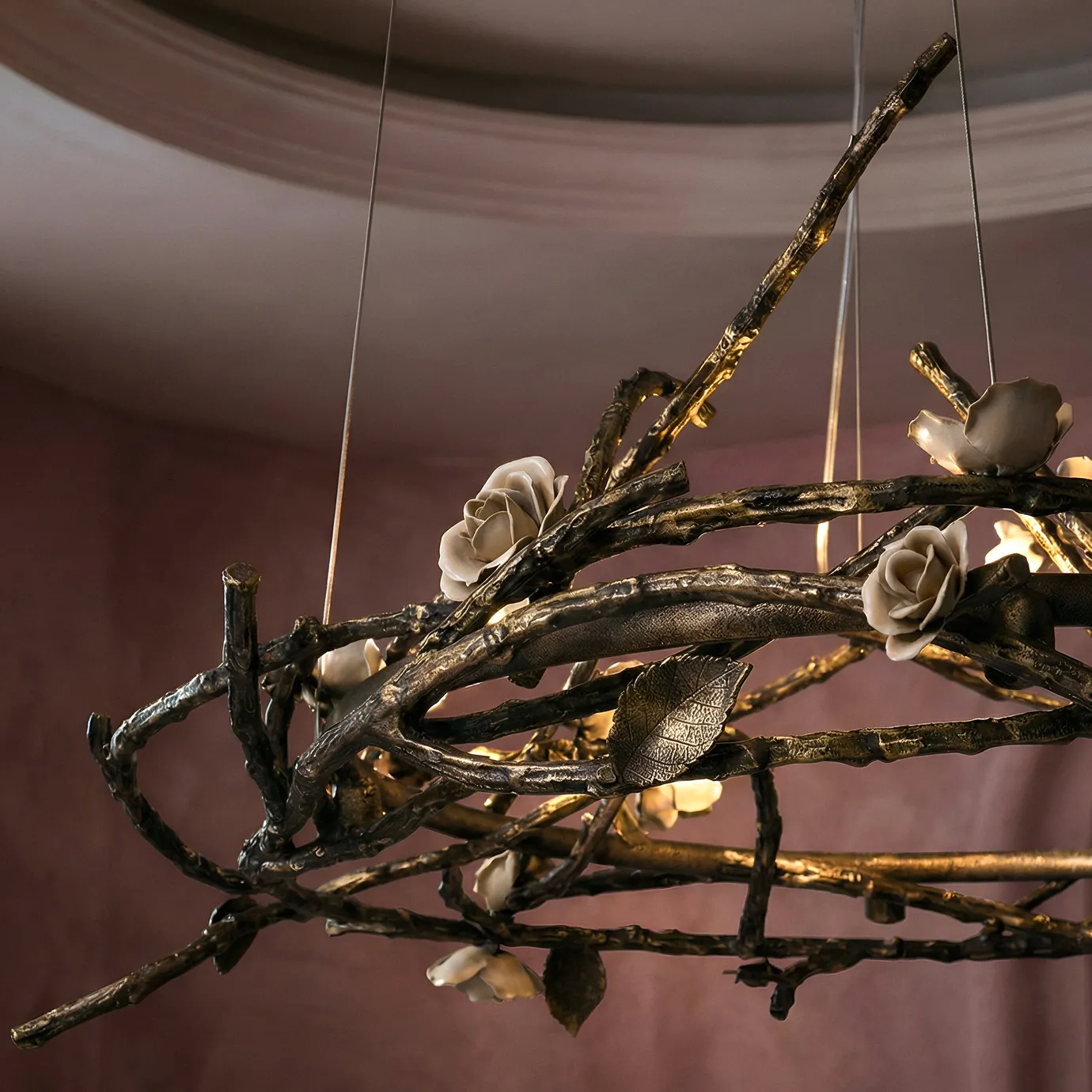 Rose Round Chandelier in aged brass with sculptural branches, metallic leaves, and cream rose-shaped accents, suspended by thin wires in a modern interior.