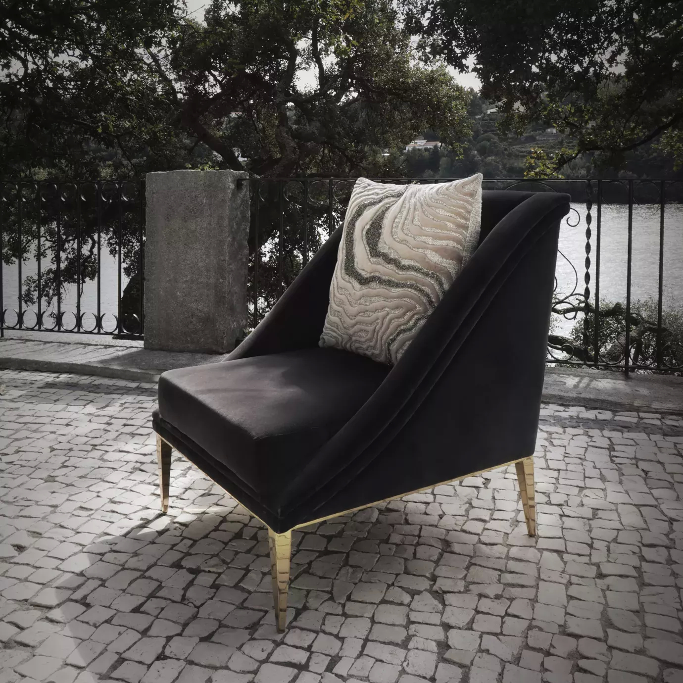 Elegant Geisha Chair upholstered in black velvet with a decorative cushion and golden legs in a sophisticated outdoor setting with a water view.