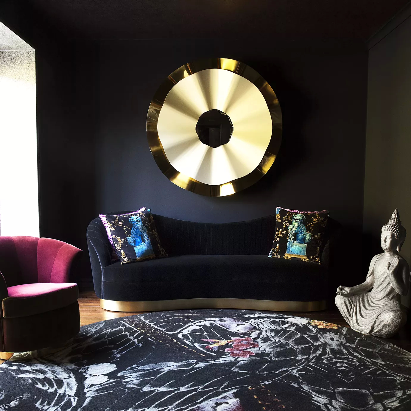 Elegant living room with a Kelly Sofa in black velvet with a golden base, decorative cushions, a burgundy armchair, and a large golden-toned mirror on a dark wall.