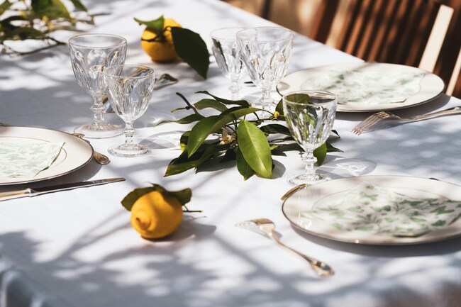 A outdoor dining space, set with a white table. There are plates, glasses and lemons on top of the table.