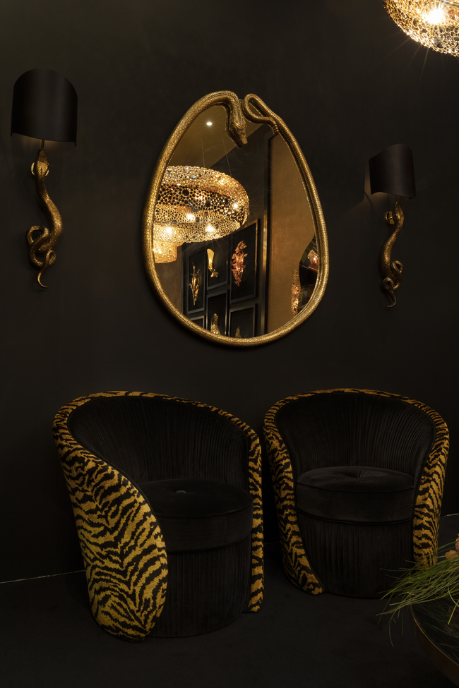An exotic room featuring two chairs upholstered in feline-print fabric. The wall is adorned with a mirror surrounded by golden snake resembling frame, accompanied by two serpent-themed sconces.