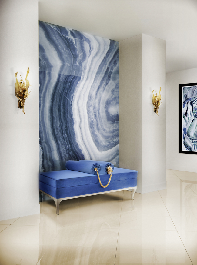 A entryway with a blue bench with two pillows. The wall behind it features a blue patterned wallpaper, centered between two golden sconces.
