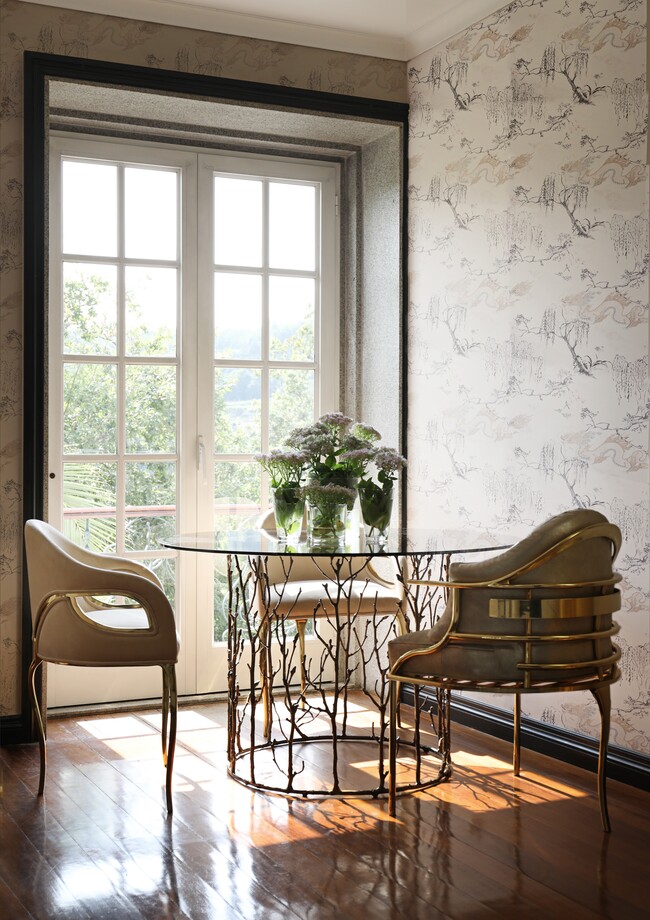 A dining room with a patterned wallpaper and a large window that lets natural light shine into the space. There is an Enchanted Dining Table, characterized by its aged brass structure resembling branches, Surrounding the table there are three chairs in cream hues.
