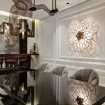 Top Interior Designers in Middle East A neutral colored room, designed by a middle east interior designer, with a glass table, three grey armchairs. Above the table here is a art structure and, on the wall, a elaborate golden sconce.