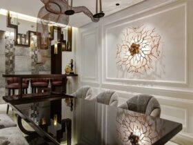 A neutral colored room, designed by a middle east interior designer, with a glass table, three grey armchairs. Above the table here is a art structure and, on the wall, a elaborate golden sconce.
