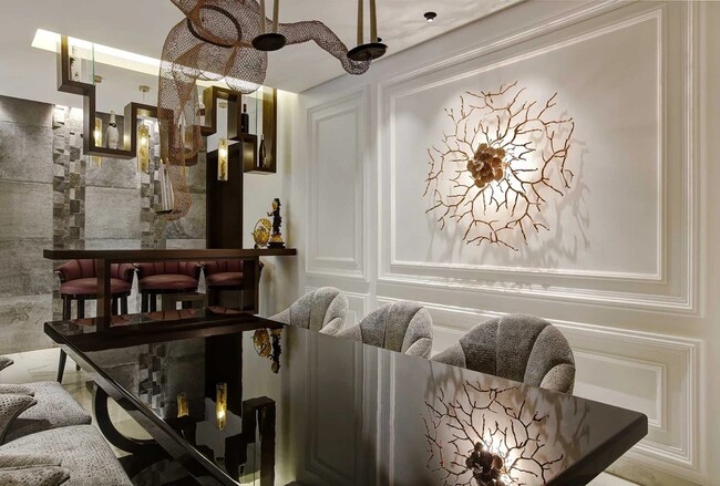 A neutral colored room, designed by a middle east interior designer, with a glass table, three grey armchairs. Above the table here is a art structure and, on the wall, a elaborate golden sconce.