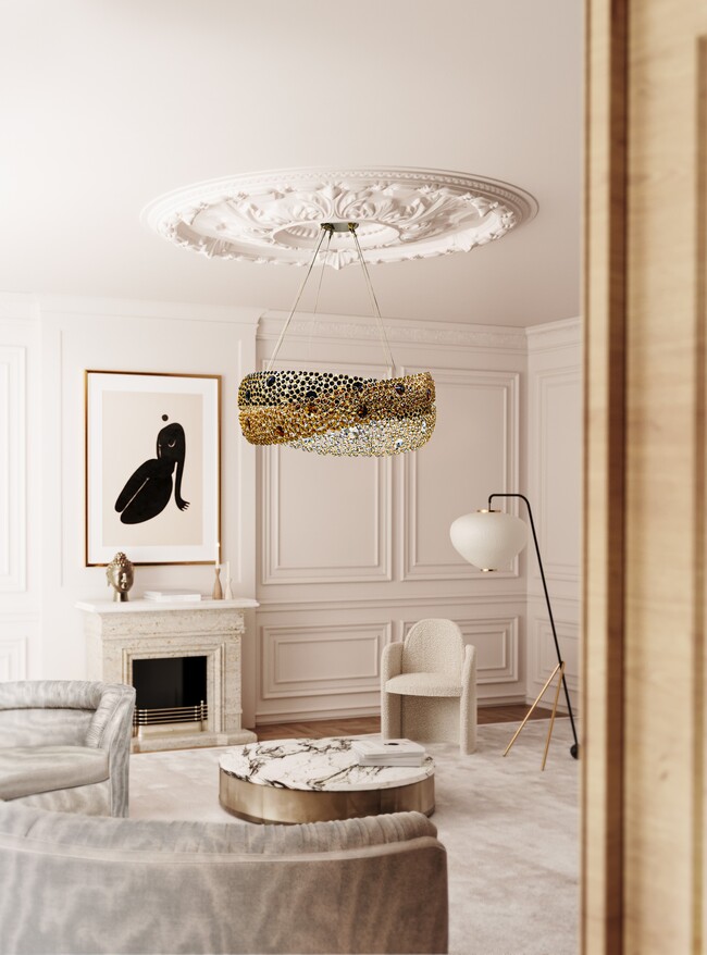 A modern living room with a fireplace decorated with a sculpture anda frame above it. The room also features a grey curved sofa, a white chair and a marble center table. Centered on the room, thre is a chandelier in golden and black hues.