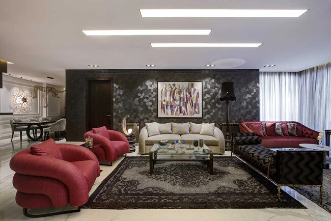 A modern living room, in dark grey and white tones, as well as a red highlight in the sofa and two armchairs. A glass table is in the center, above a rug. On the background, a painting is hung on the wall.
