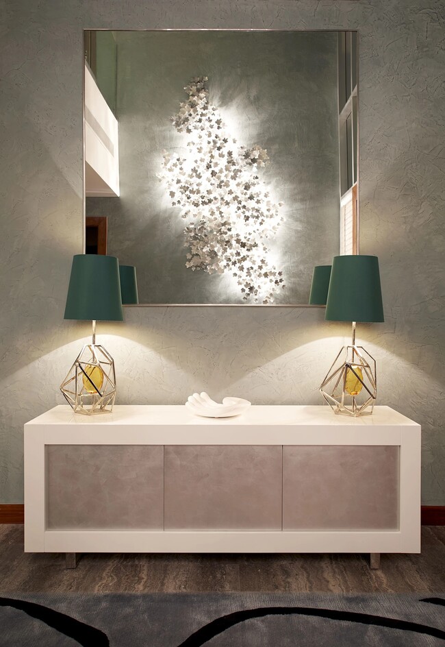 A entryway in neutral colors, with a big mirror and a minimal white cabinet. On the cabinet, there are two table lamps, in golden and dark green shades.