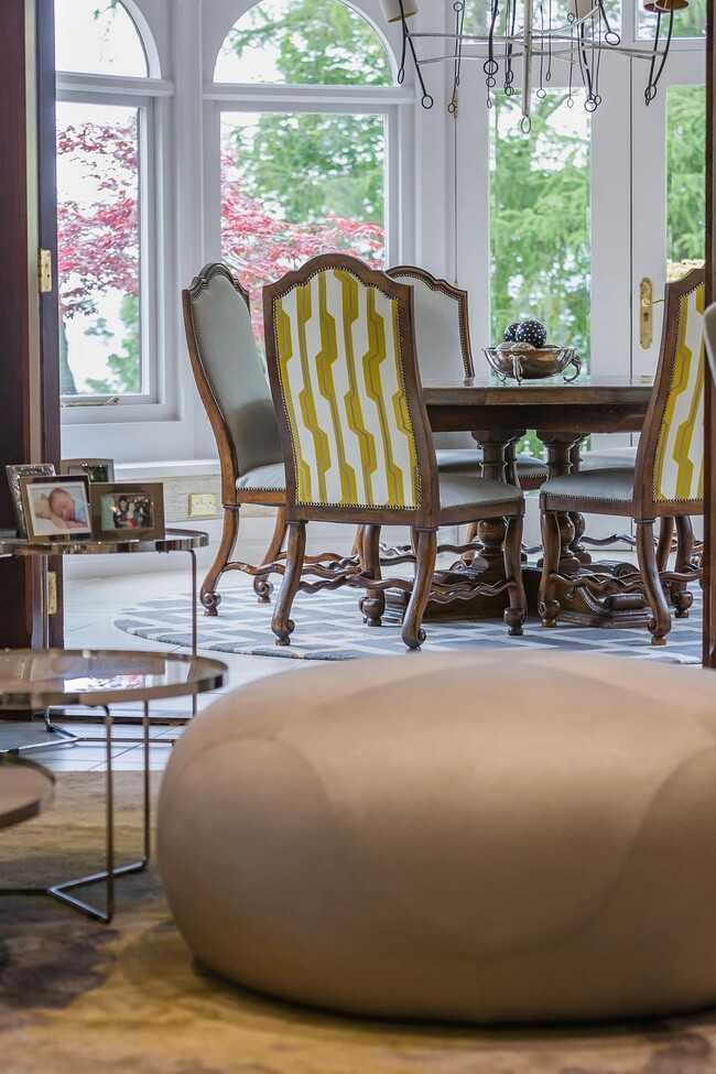 A dining room in earthy tones, with a wooden dining table, surrounded by four wood chairs with white and yellow upholstery. In the background there are large windows.