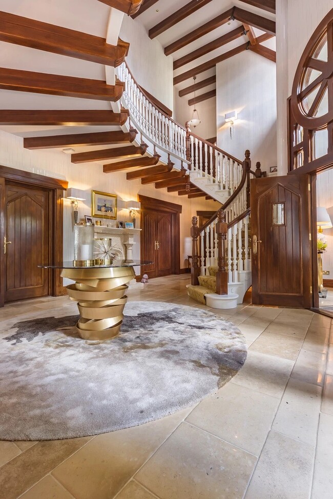 A large entryway, in tones of white and brown. The focal point of this space is the Intuition Center Table, with a glass tabletop and a glamourous golden swirling structure, placed on a rug. In the background, there is a staircase and big wood doors.