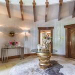 An airy entryway in neutral tones. The focal point of this space is the Intuition Center Table, with a glass tabletop and a glamourous golden swirling structure, placed on a rug. Next to the center table, there is a mirror and table. In the background, a wooden door is seen.