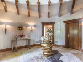 An airy entryway in neutral tones. The focal point of this space is the Intuition Center Table, with a glass tabletop and a glamourous golden swirling structure, placed on a rug. Next to the center table, there is a mirror and table. In the background, a wooden door is seen.