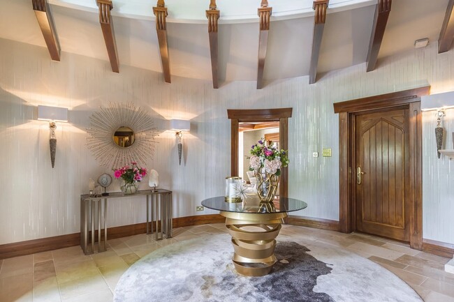 An airy entryway in neutral tones. The focal point of this space is the Intuition Center Table, with a glass tabletop and a glamourous golden swirling structure, placed on a rug. Next to the center table, there is a mirror and table. In the background, a wooden door is seen.