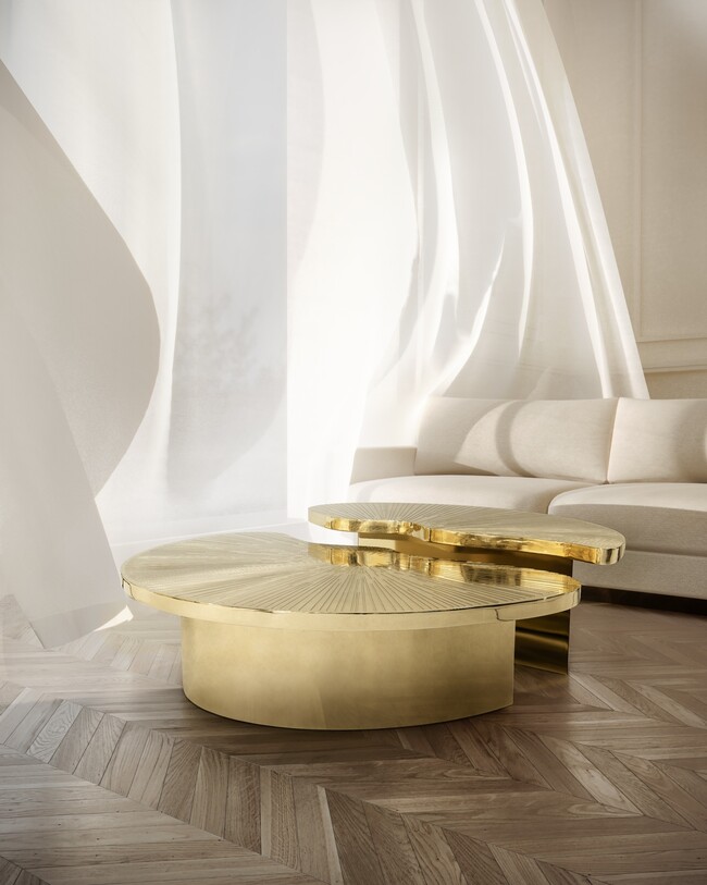 A neutral colored living room with a cream sofa and a golden center table, divided in two different heighted parts. A window is open, letting the breese flow the white curtains into the space.