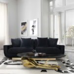 A modern and luxurious living room, in neutral colors. In the center of the room there is a black sofa, combining with the elegant center table, that serves a statement piece as it resembles the legs of a woman. In the background, there are frames with visual art.