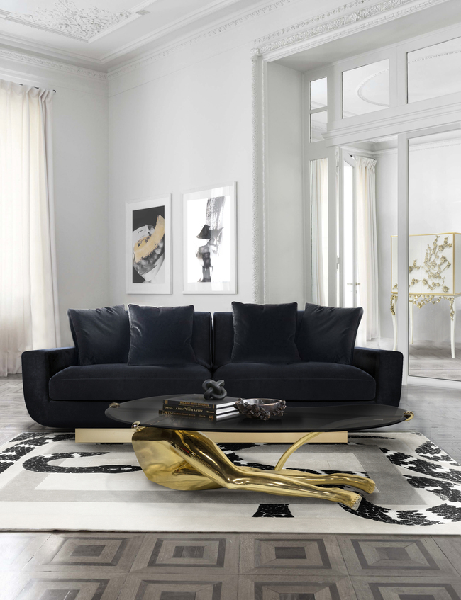 A modern and luxurious living room, in neutral colors. In the center of the room there is a black sofa, combining with the elegant center table, that serves a statement piece as it resembles the legs of a woman. In the background, there are frames with visual art.